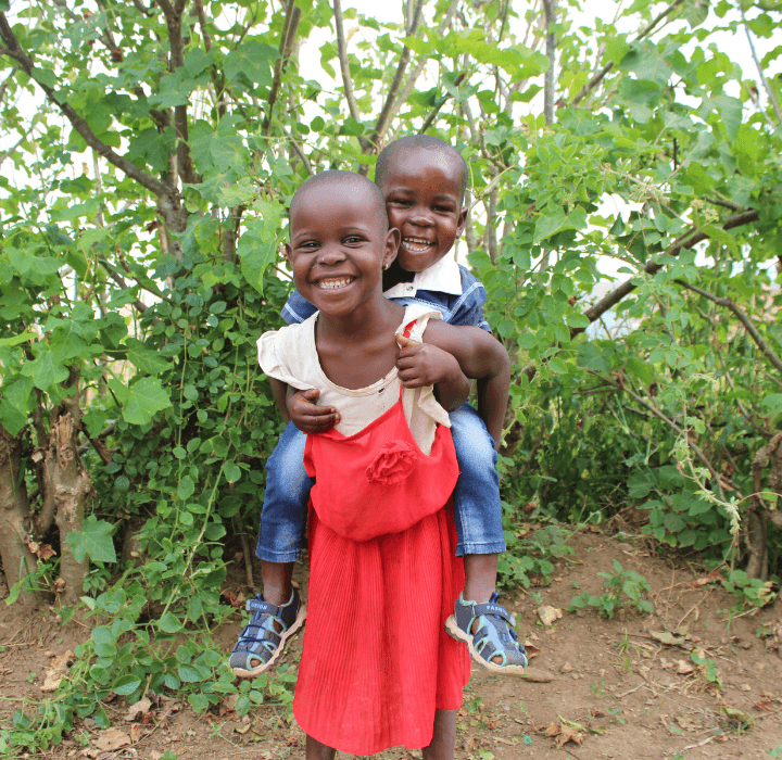 Two young foster siblings in LWB's Uganda foster care program smiles big at the camera. The older girl in red is giving the younger boy in blue a piggyback ride