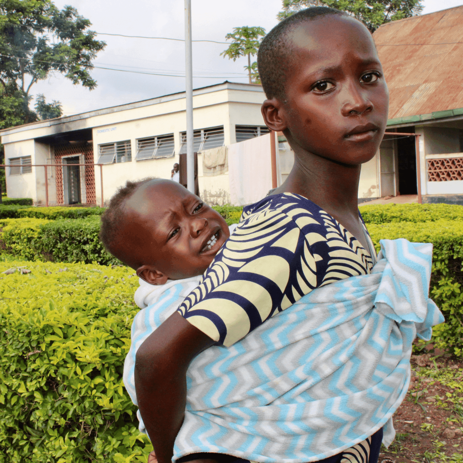 A young Ugandan mother holds her sick and crying baby in a blanket on her back.