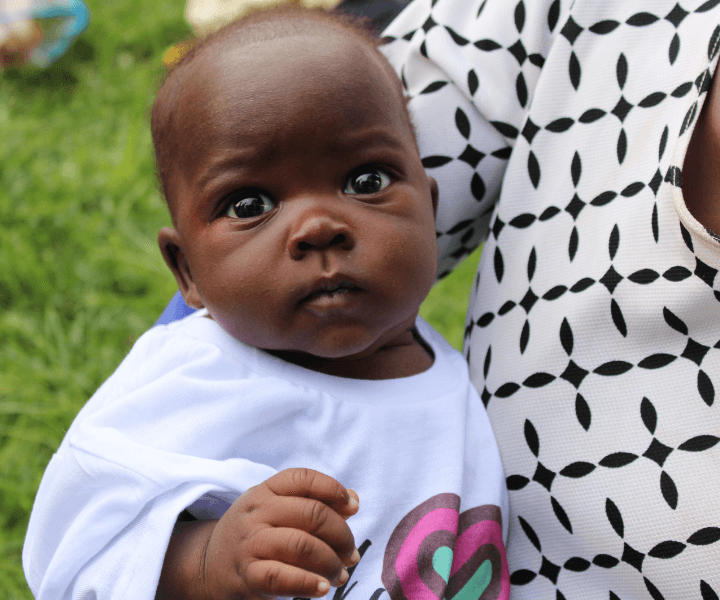 A Ugandan baby in an LWB white shirt is being held by his mother.