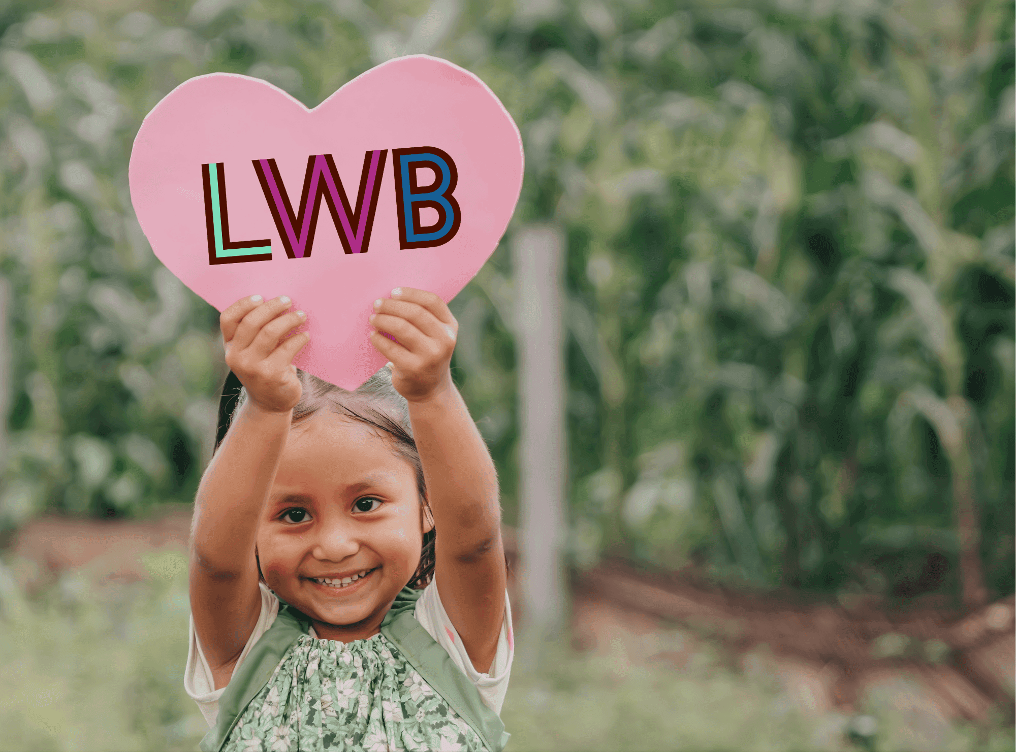 Guatemalan Girl Holding up a Pink Heart with LWB Logo on it. She stands outdoors in front of a field of corn.