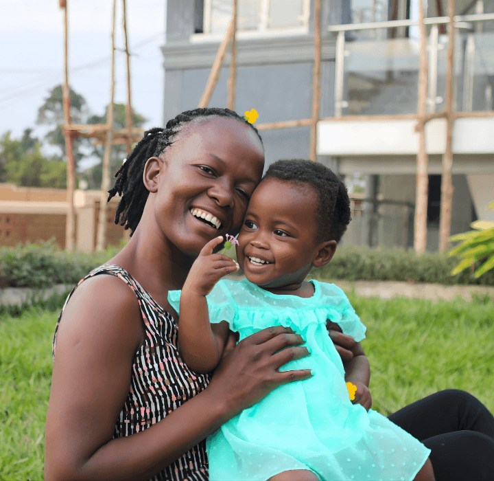 A toddler girl from Uganda sits on the lap of her foster mother who holds her close. They are both smiling, while the little girl holds a flower in each hand.