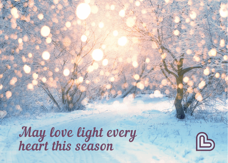 The woods with snowy ground and the light reflecting off the falling snow like twinkle lights. The message says, "May love light every heart this season."