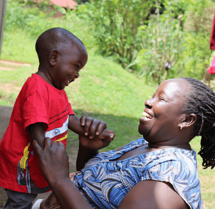 A toddler boy from Uganda is laughing with his foster mom who loving holds on to his arms. She sits at his level so that they are looking into each others' eyes