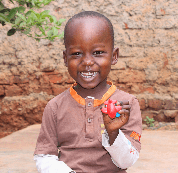 A little Ugandan boy holds up a red airplane toy. His big smile shows his bright teeth. He wears a brown shirt with an orange collar.