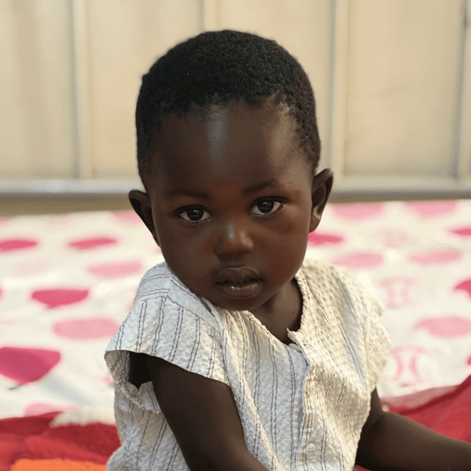 A toddler-aged Ugandan girl stares intently at the camera. She sits on a pink and white polka dot blanket.