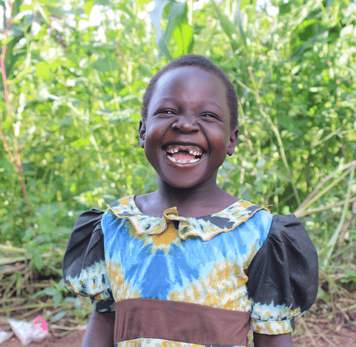 A young Ugandan girl with short hair laughs heartily, showing that her two front teeth are missing. She wears a blue and yellow dress with tie-dyed patterns.