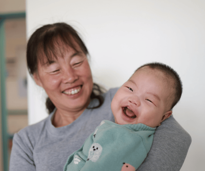 Smiling mother holding her baby boy, who is laughing with his new smile after his corrective cleft lip surgery.