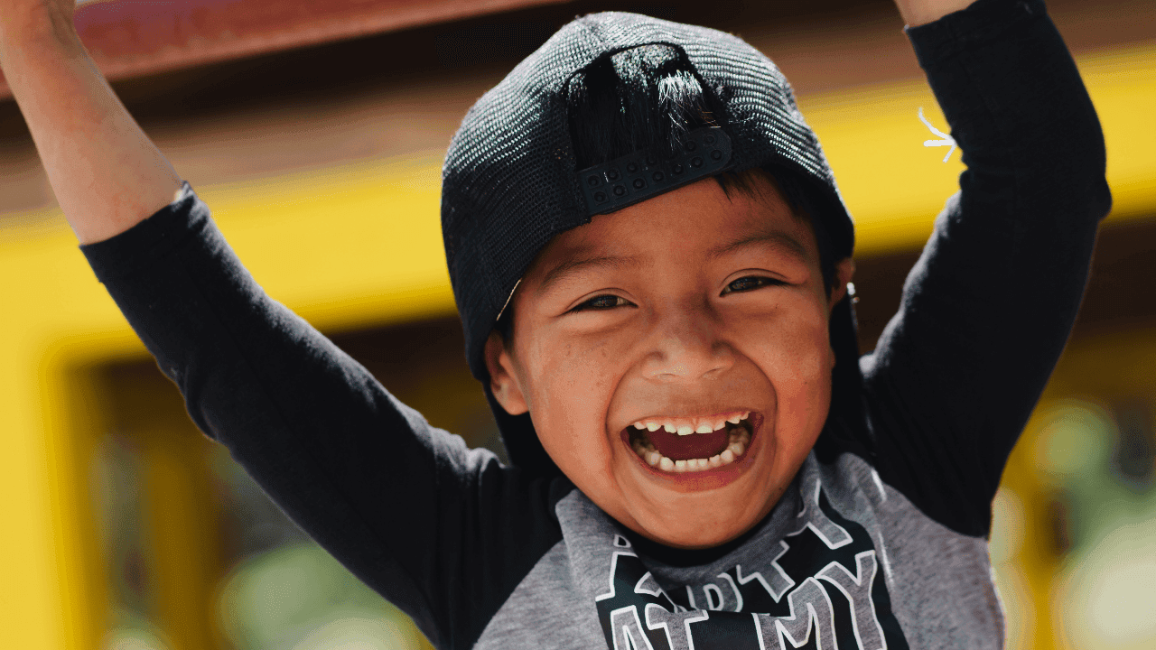 A boy in LWB's Education Program hands on the jungle gym with a big smile. A navy cap sits backwards on top of his head, and he wears a gray and navy shirt.