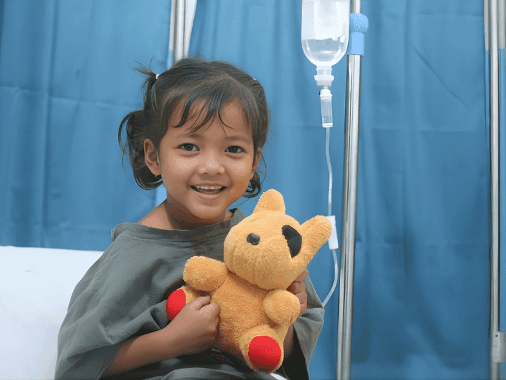 A young Indian girl smiles with her stuffed animal. She sits on a hospital bed in a hospital gown with an IV drip. Behind her is a blue curtain.