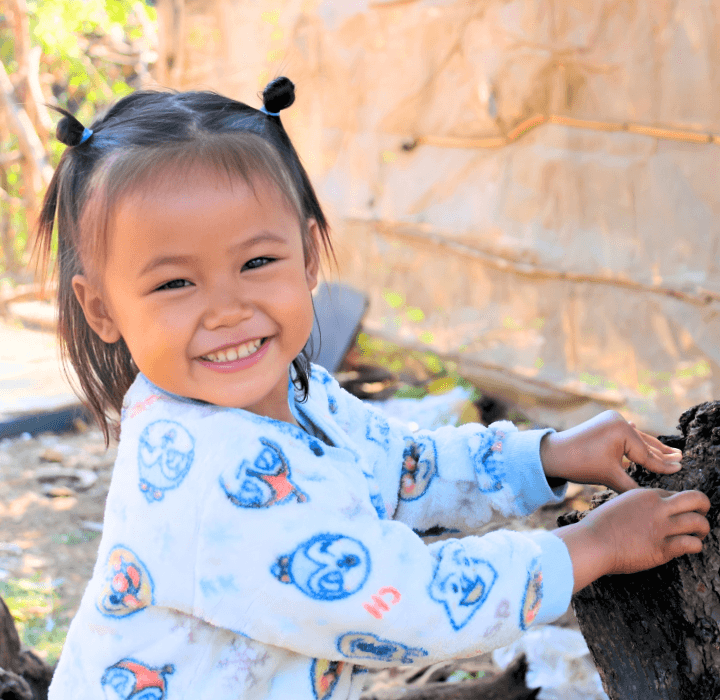 A young Cambodian girl with two hair buns on her head sits beside a tree trunk. She smiles brightly and wears a white set of pajamas with cartoon faces on it.