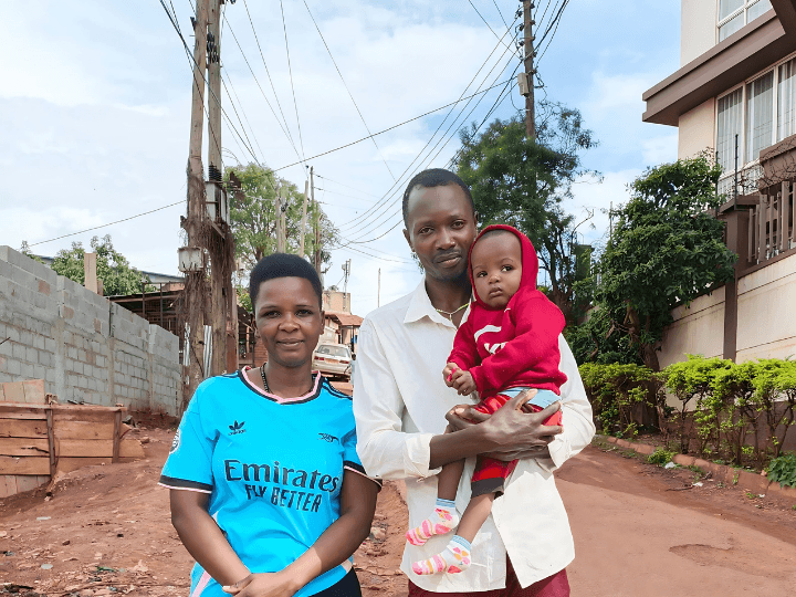 A mother and father stand in a Ugandan street holding their toddler son. The boy wears a red sweater with his hood up. The mom is in blue; the father in white.