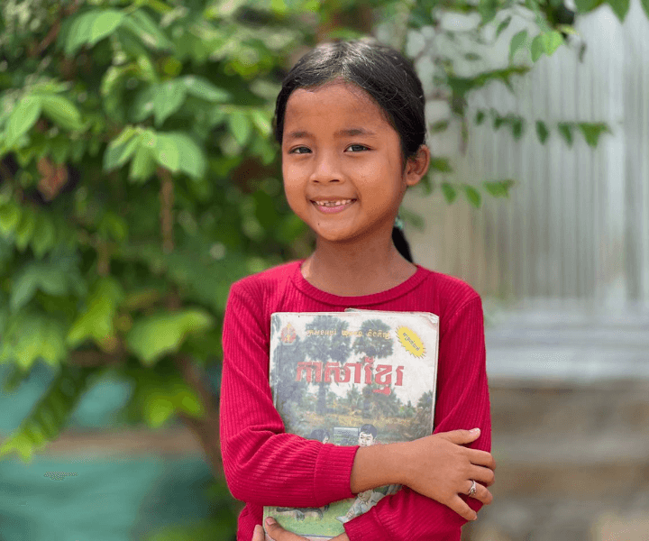 School-aged girl smiles and holds a Khmer book to her chest.