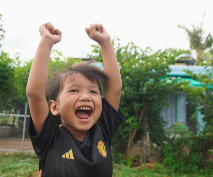 A little girl in Cambodia pumps her two fists up in the air in excitement. He wears an LWB Believe in Me School uniform.
