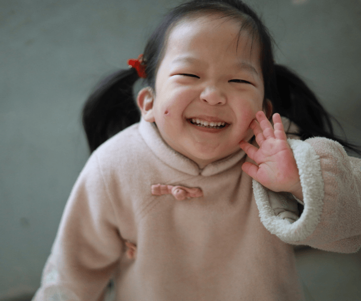 A girl from LWB's healing home in China smiles broadly at the camera. She wears a traditionally styled soft pink fleece sweater.