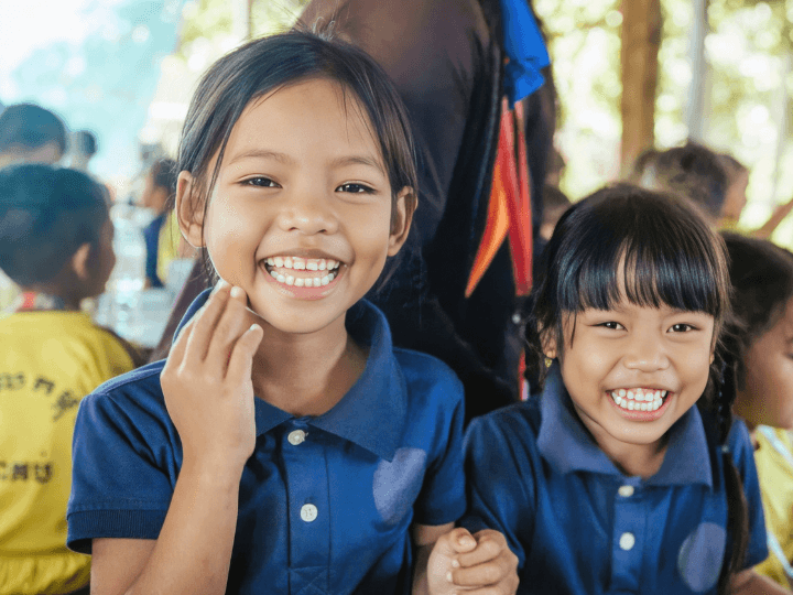 Two Cambodian girls in their blue school uniform sit together in a crowded covered outdoor sitting area. They laugh with big smiles while looking at the camera.