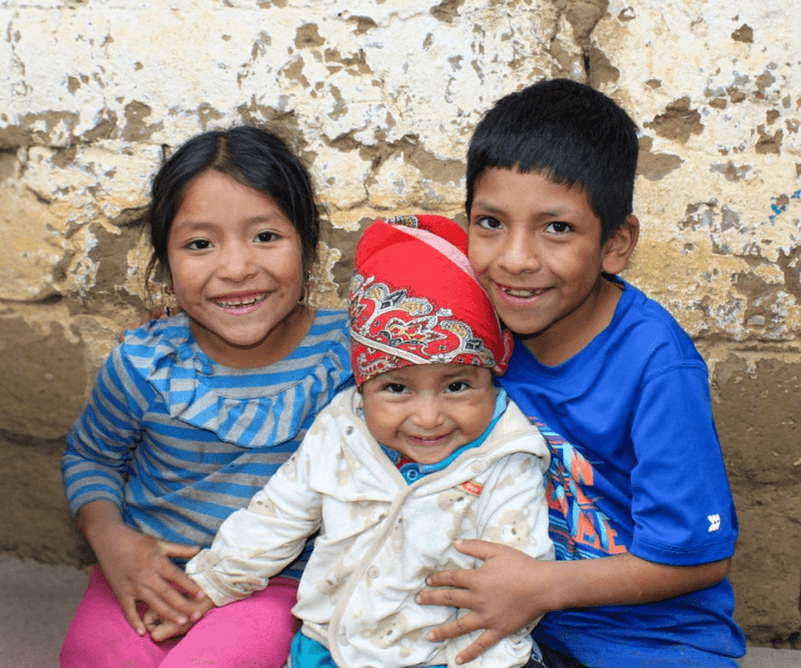 A school-aged brother and sister hold their toddler-aged sister while sitting on a bench. They all smile broadly at LWB's Tob'Anik Education Center in Guatemala
