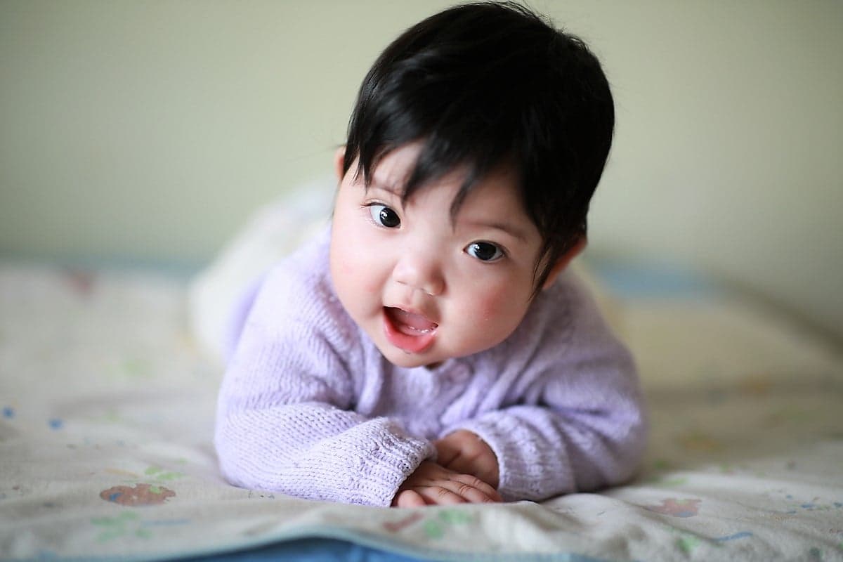A Chinese baby girl with a full head of black hair and wearing a purple knitted sweater lays on her belly on a bed.