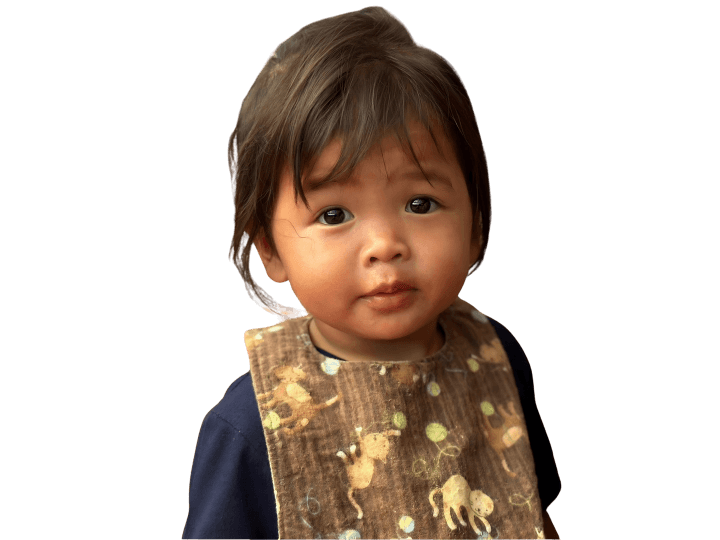A preschool-aged Cambodian girl looks hopefully at the camera with her big eyes. She wears a brown bib with cats on it.