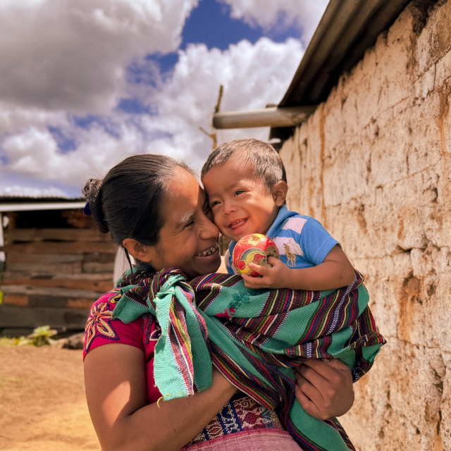 A toddler laughs in his mother's arms as he holds a ball in his hand.