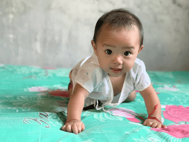 A Chinese baby is on a bed covered with a teal blanket with designs of red roses. He crawls toward and looks at the camera. He wears a red thread on his wrist.