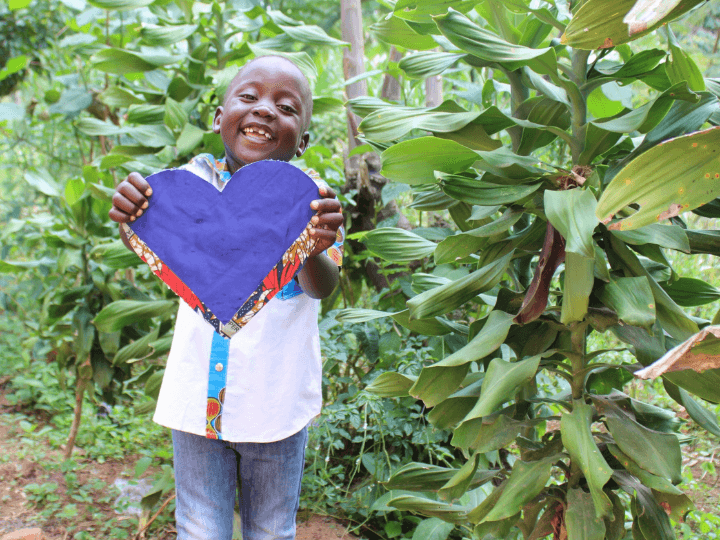 An elementary-aged Ugandan boy stands outdoors amongst trees. He smiles with a front tooth missing and holds a large heart crafted from purple paper.