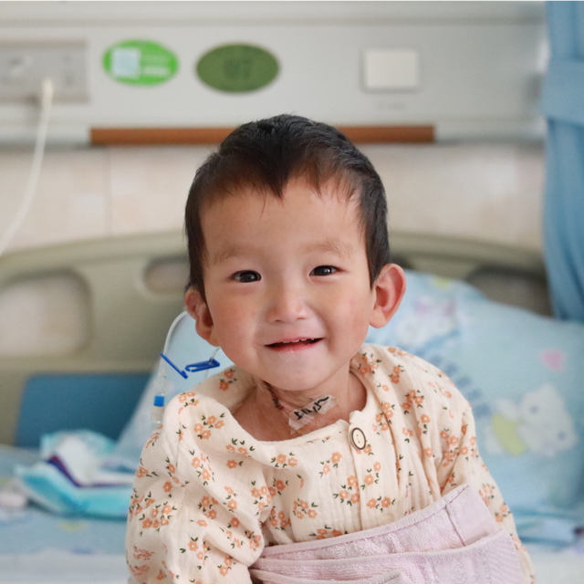 A toddler girl smiles happily while sitting on a hospital bed.
