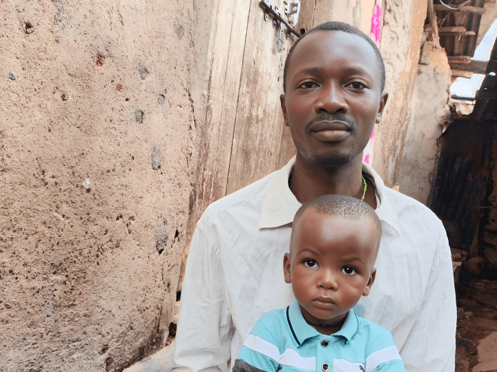 A little boy and his dad from Uganda sit together in front of a rock wall with a wooden gate. The father is in white. The boy wears a blue and white polo shirt.
