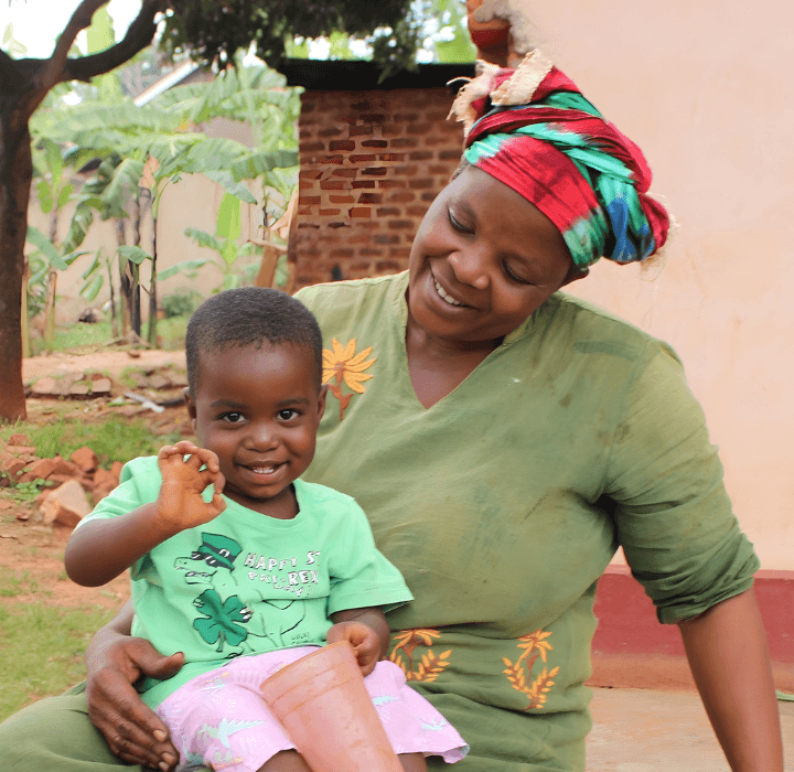 A preschool-aged boy sits outdoors in the lap of his foster mother who smiles at him adoringly. He smiles and looks at the camera and holds one of his hands up.