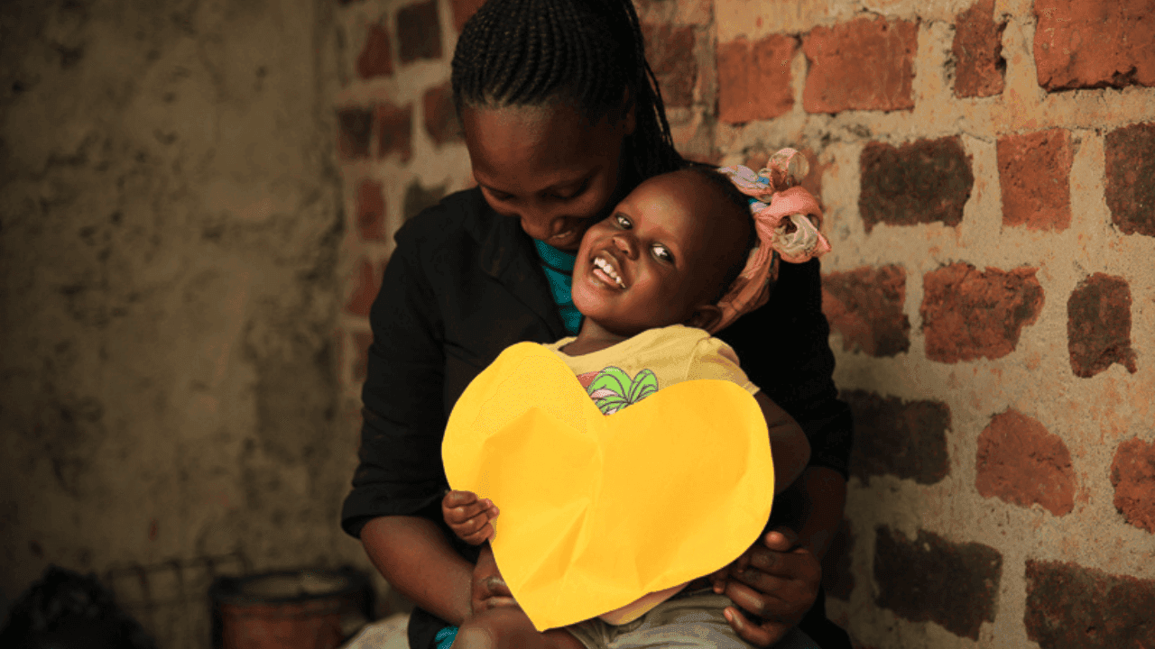 A mother and daughter from Uganda smile while sitting in front of a brick wall. The young girl holds a large yellow paper cutout shaped like a heart.