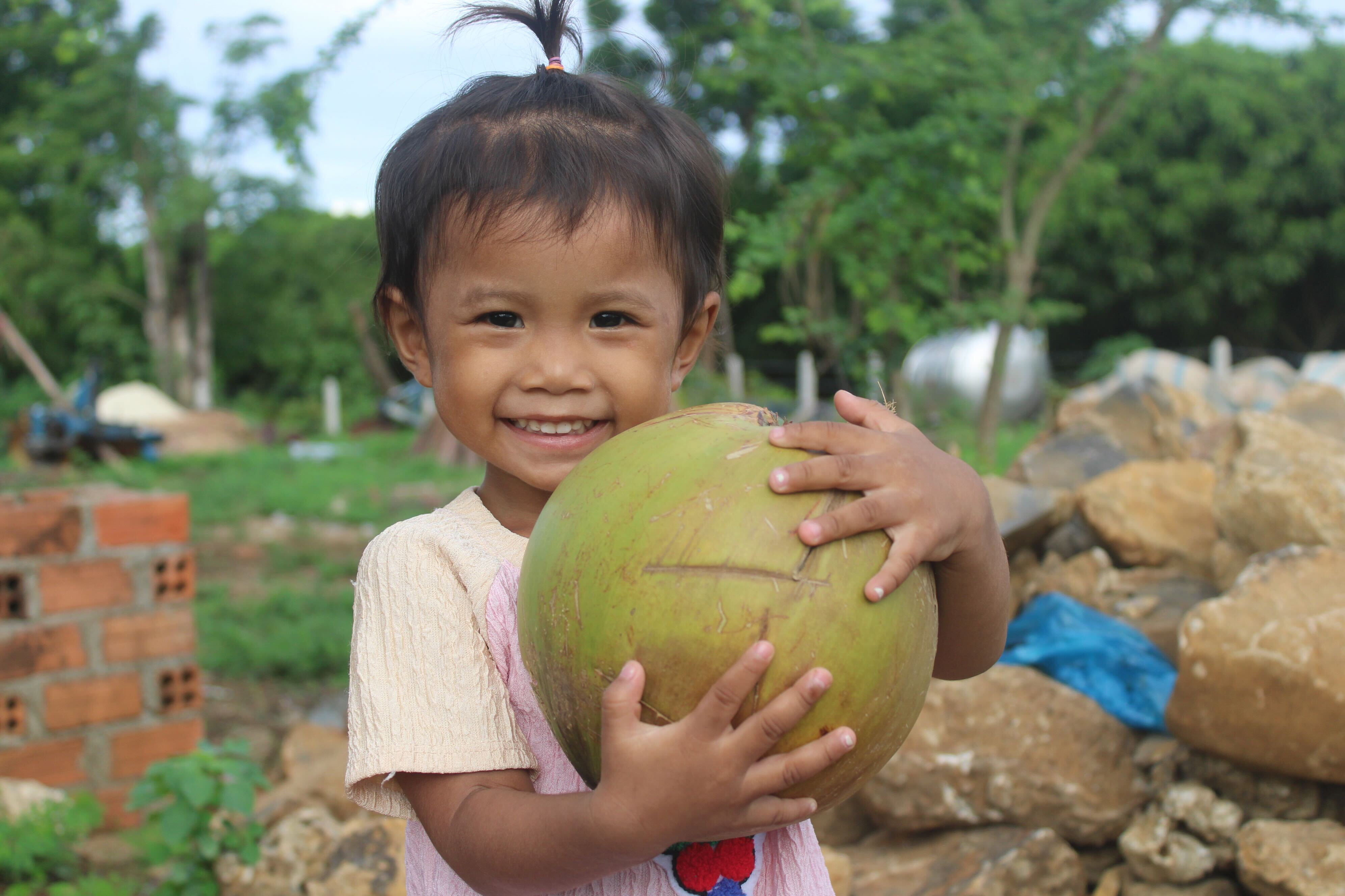 An elementary-aged Cambodian girl with a pigtail smiles widely while holding a giant coconut the size of her head. 