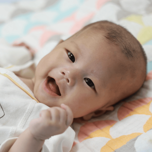 A smiling Chinese baby is lying on a beautiful quilt.