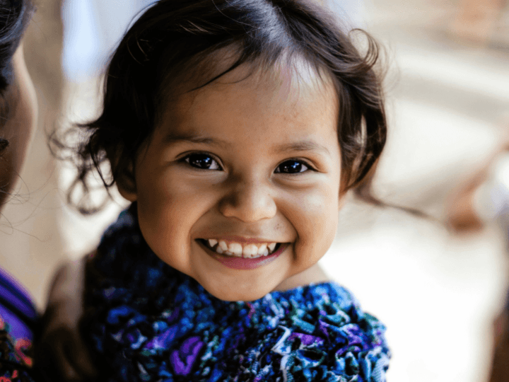 A close up of a toddler girl from Guatemala. She wears a big smile and a purple, blue, and teal shirt. Her eyes shine bright at the camera.