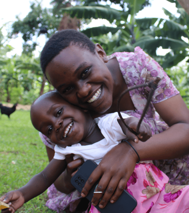 A girl in LWB's Uganda Foster Care Program laughs as her foster mom holds her and hugs her from behind. They both wear pink and purple dresses and smile widely.