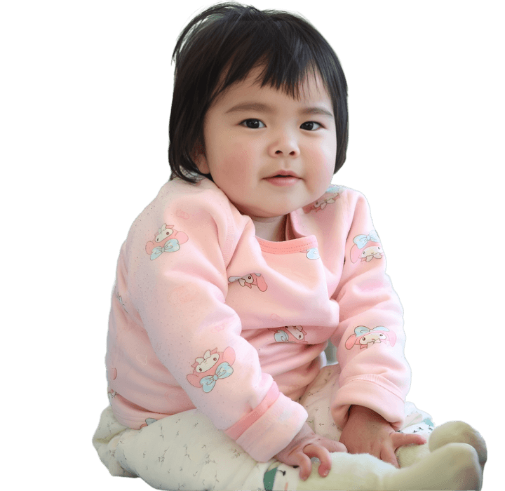 A Chinese toddler girl sits on a hospital bed. She smiles at the camera with her hands neatly on her lap. She wears a pink sweater, white pants, and white socks