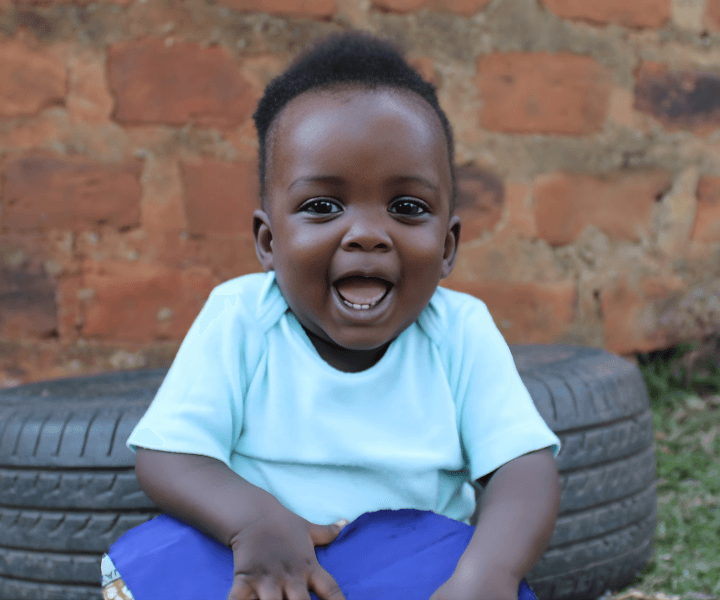 Ugandan boy smiles as he holds a purple heart.  He sits outdoors in front of a brick wall and a tire.