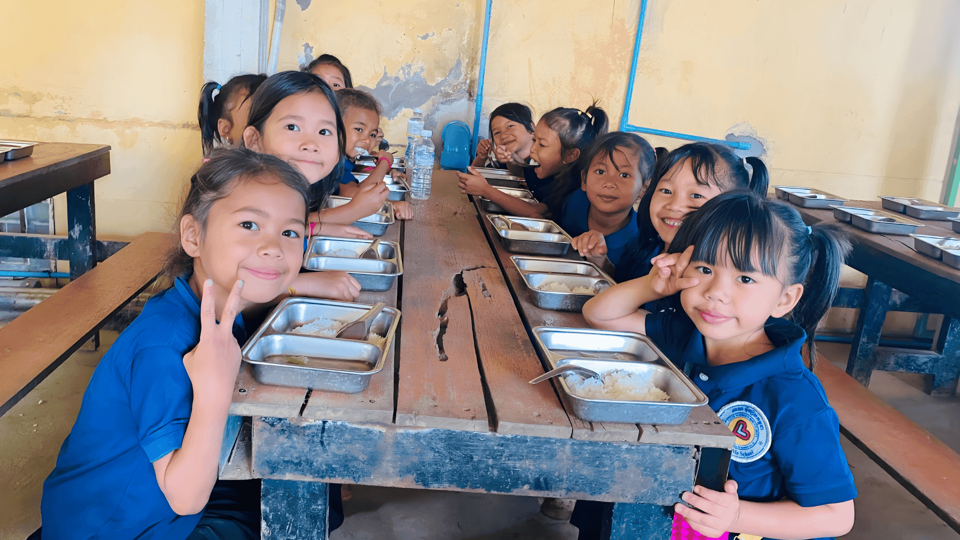 A group of young Cambodian girls in LWB's blue school uniforms fill both sides of a lunch table, eating rice and a soup containing meat and vegetables. 