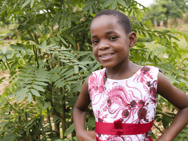 A Ugandan school-aged girl stands in front of a tree. She smiles and wears a white dress with an intricate pattern of red beads and a red sash at the waist