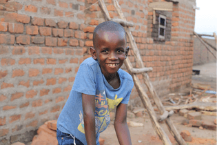 A young boy in LWB's program stands in front of a brick wall and wooden ladder. He wears a blue shirt and jeans. He smiles brightly at the camera.