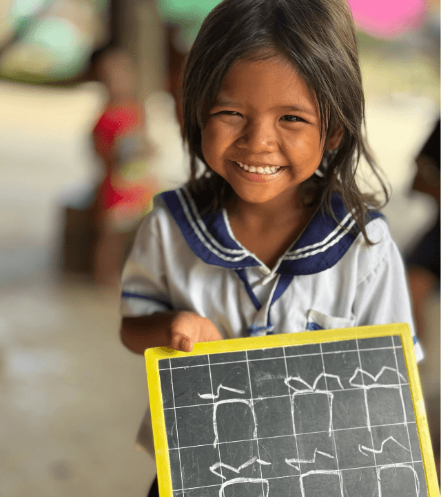 A smiling Cambodian girl holding a chalkboard with her Khmer characters written on it.