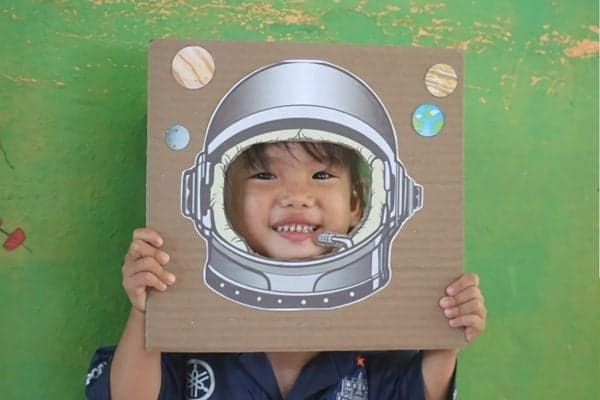 A young girl smiles and holds a large cardboard prop of an astronaut helmet up to her face. The prop has a hole so the girl's face fits inside the visor area.