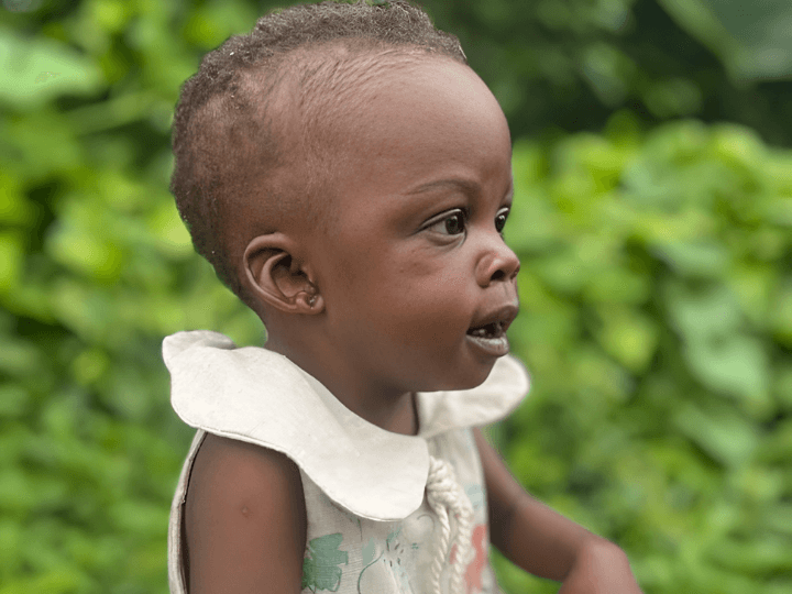 A skinny little Ugandan girl around the age of one years old looks to her left. She wears a white dress. Behind her are green bushes.
