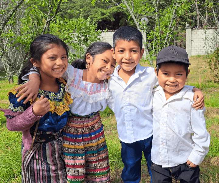 Four school-aged kids in traditional Guatemala clothing are in a group hug. They laugh and smile at the camera.