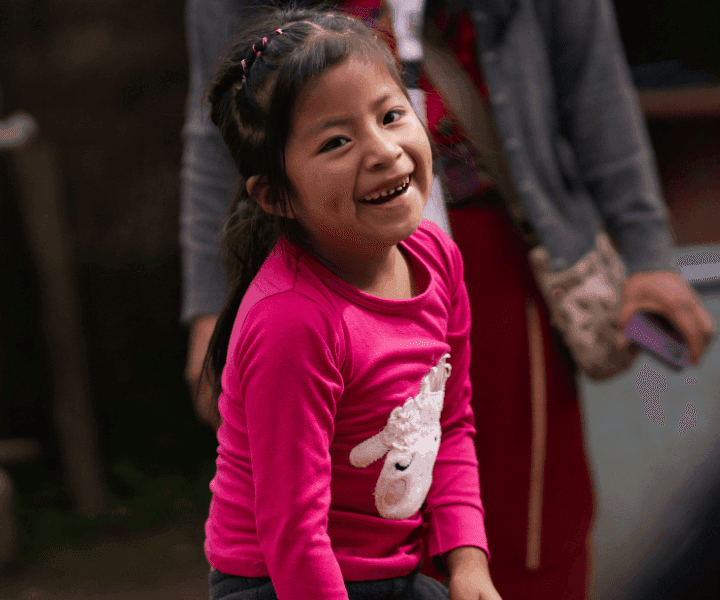A girl in a bright pink shirt with a picture of a lamb on it smiles with dimples on her cheeks. Her hair is in a French braid.