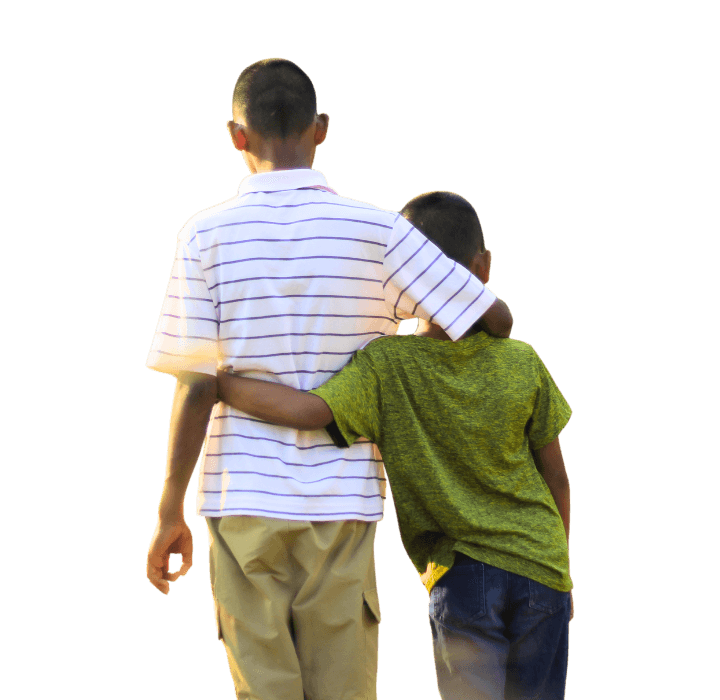 Two brothers walk away from the camera one arm around the other. They walk in a field of harvested wheat.